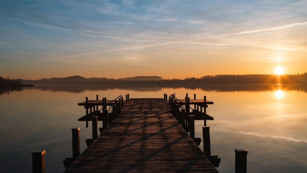 A long wooden dock extends into a calm lake toward a rising sun on the horizon.