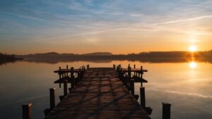 A long wooden dock extends into a calm lake toward a rising sun on the horizon.