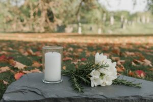 A white candle in a glass holder sits beside small white freesias and rosemary on a dark stone slab outdoors among fallen autumn leaves, with blurred gravestones in the background.